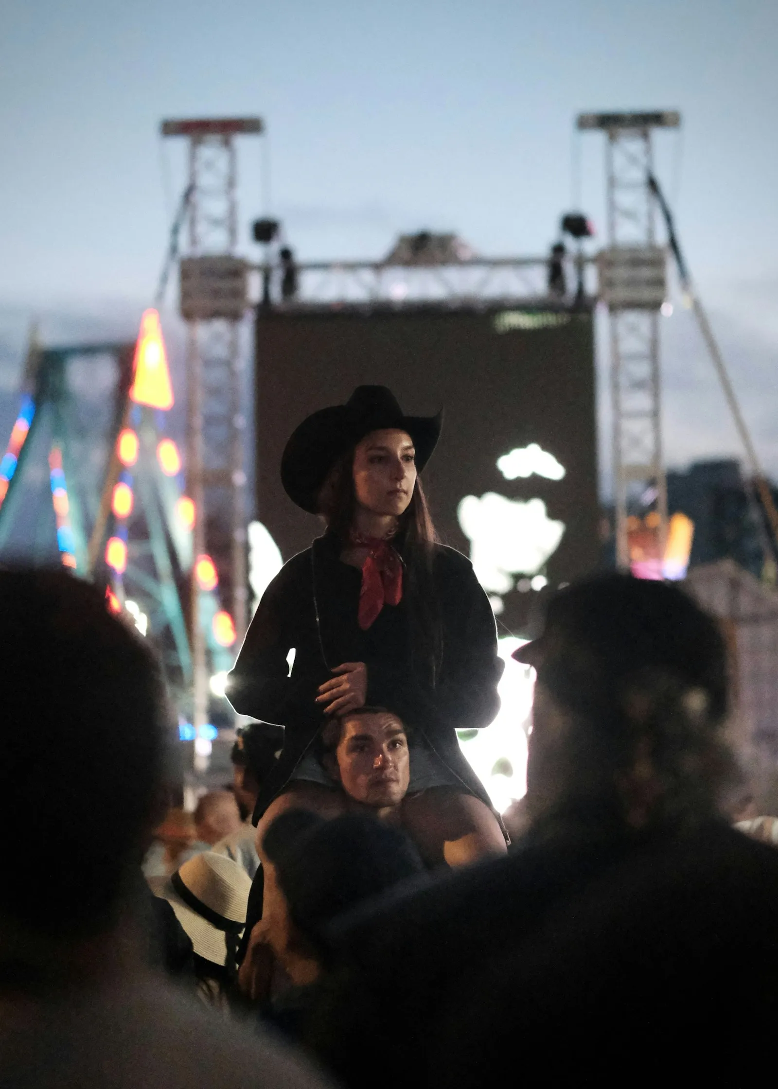 Žena v černém kovbojském klobouku sedí někomu na ramenou v davu na festivalu za soumraku
